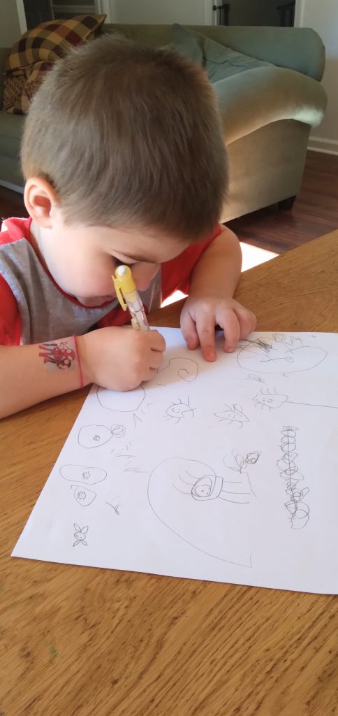 Young brown-haired boy sitting at a table drawing spiders with a yellow pencil.
