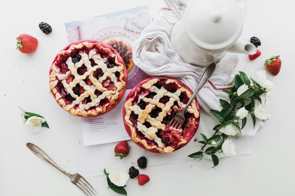 White counter covered with stray pieces of strawberry and raspberry fruit, a white hand towel, white flowers, white tea pot and two strawberry raspberry pies.  One partially eaten.