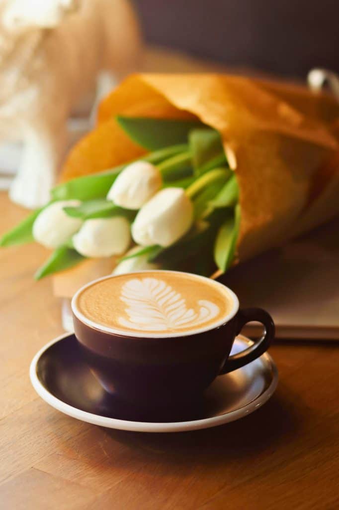 Close up of a black coffee mug and saucer with a fun floral design made from milk in the coffee.  White tulips wrapped in brown paper in the background.