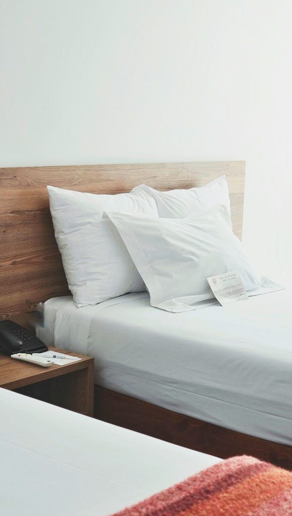 Neatly made twin size hotel bed with white sheets against a wooden headboard. Beside the bed is a wooden end table with a black telephone, white notepad and pen and white remote sitting on top.