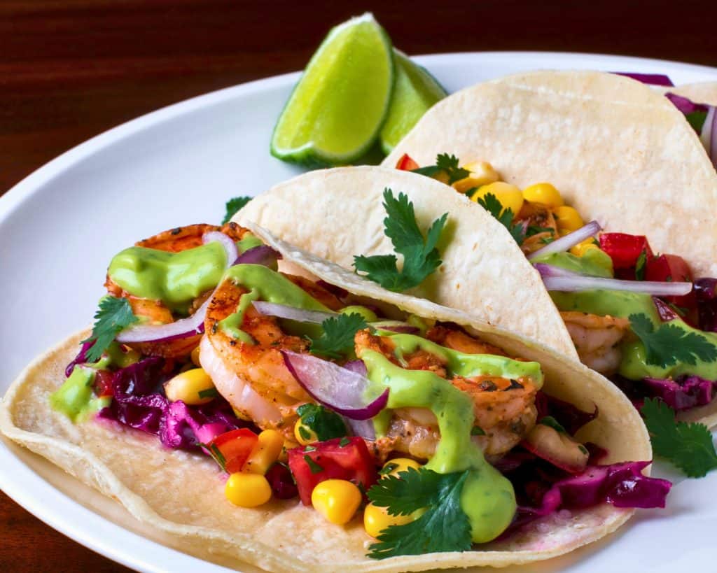 Close up of a shrimp taco in a flour tortilla topped with corn, tomatoes, purple onions, cilantros, and a green sauce.