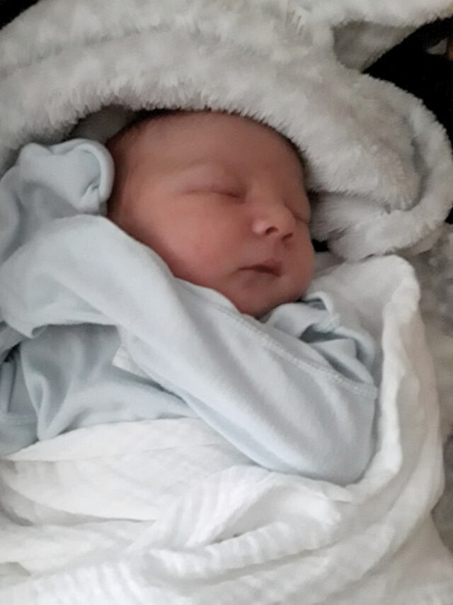Baby sleeping in a blue gown and surrounded by a grey fluffy blanket.