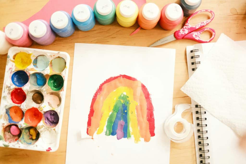 A child painting of a rainbow sitting on a wooden table surrounded by various paint colors, red child scissors, tape, and a spiral notebook.