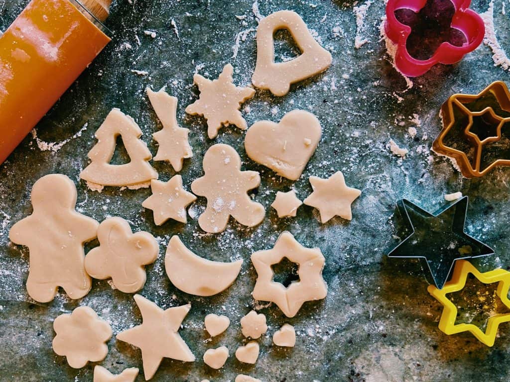 Floured counter filled with Holiday Cookie Cutters, uncooked sugar cookie dough cut in various shapes, and a rolling pin.