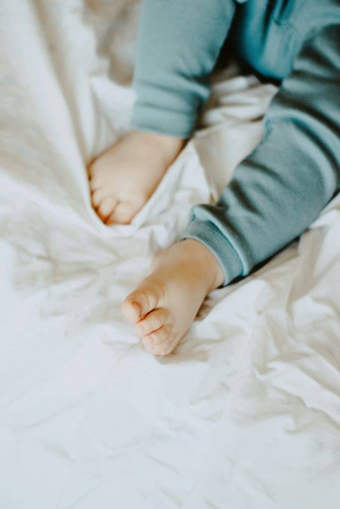 Toddler legs, wearing light blue pjs, and feet on a bed with white sheets.