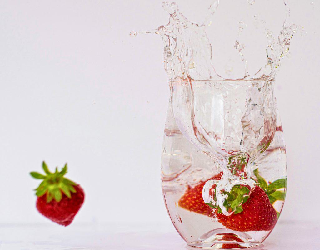 Glass cup filled with water and strawberries being dropped into it. 