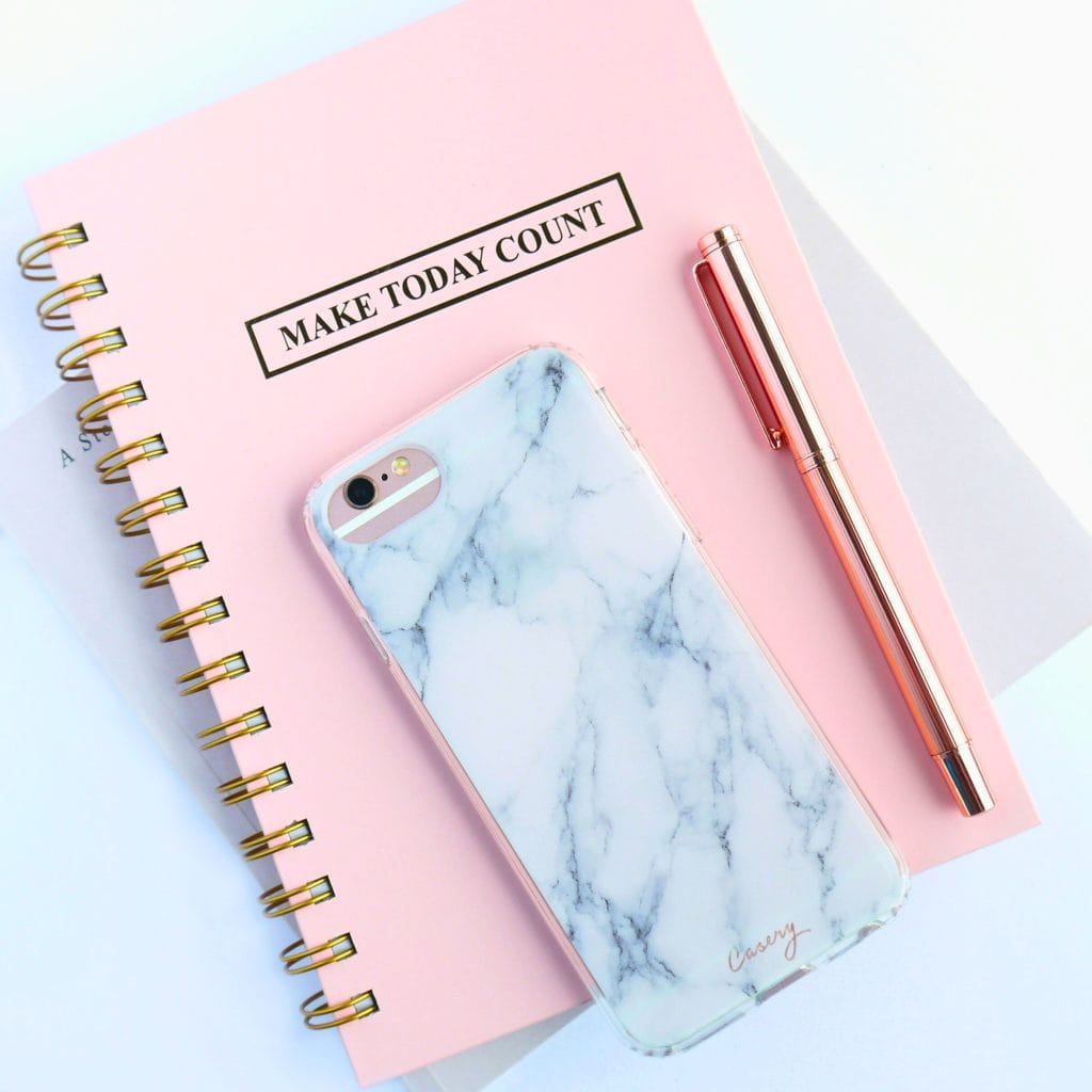 Pink spiral notebook with a rose gold pen, and marble colored cell phone resting on top.