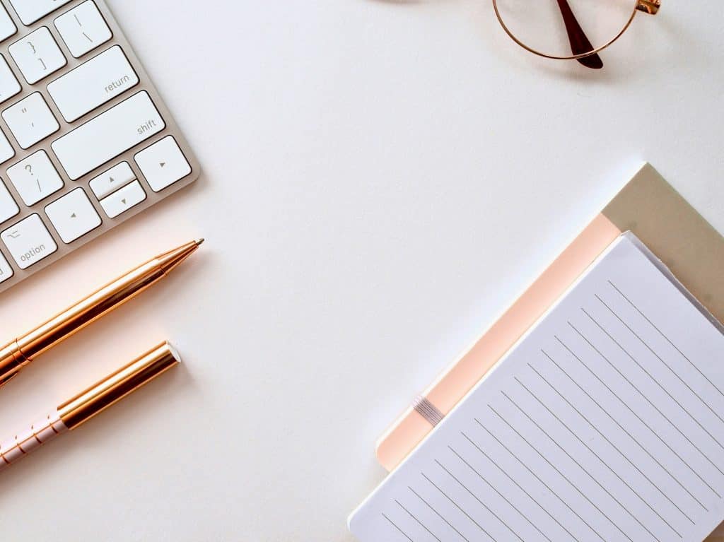 White desk surface with a keyboard, gold pens, pair of glasses, and a pink notebook sitting on top.