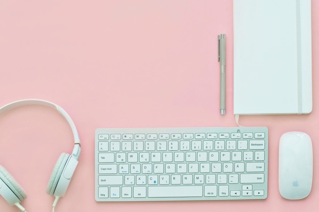 White notebook, and grey pen sitting on a pink background. Underneath those items are a white computer mouse, white keyboard, and white headphones.