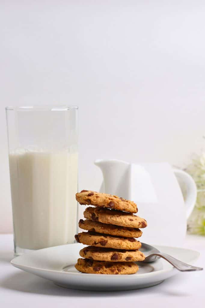 Six chocolate chip cookies on a white plate with a glass of milk and white pitcher in the background.