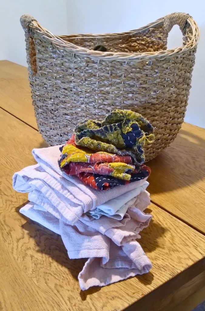 Wicker laundry basket sitting behind kitchen hand towels of various colors folded and stacked in a pile.
