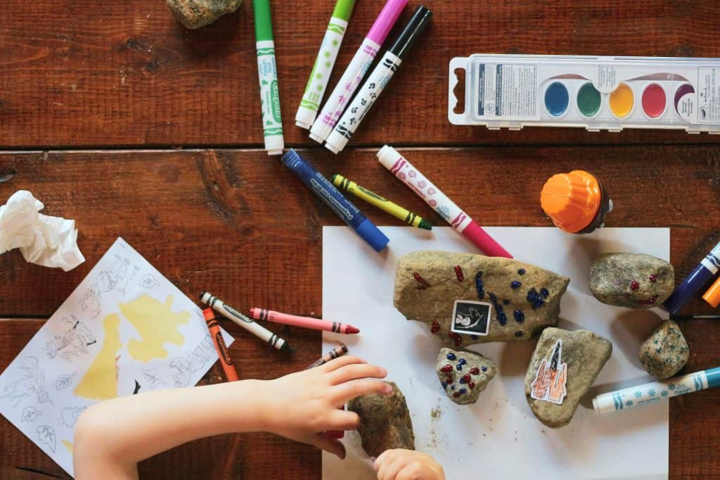 Wooden table covered with assorted markers, watercolors, crayons, rocks, and white paper. While a child colors a rock.