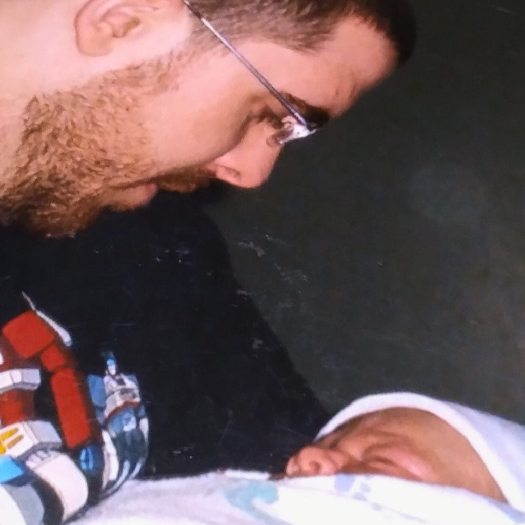 Father wearing glasses gazing down at his new baby wearing a white hat and wrapped in a swaddling blanket.