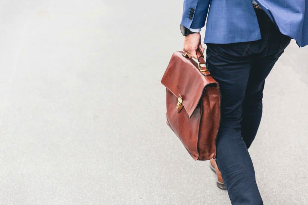 Man wearing a light blue sport coat, solid thick black watch, and navy dress pants carrying a leather briefcase.