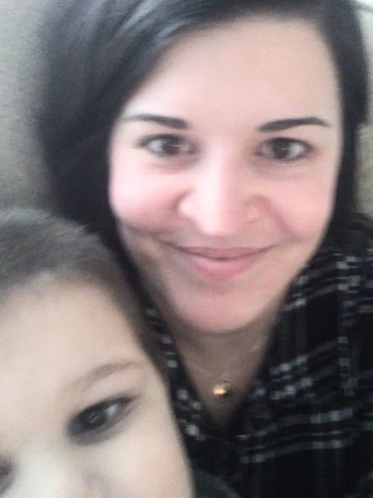 Woman with dark hair smiling at the camera with a young boy peeking into the camera.