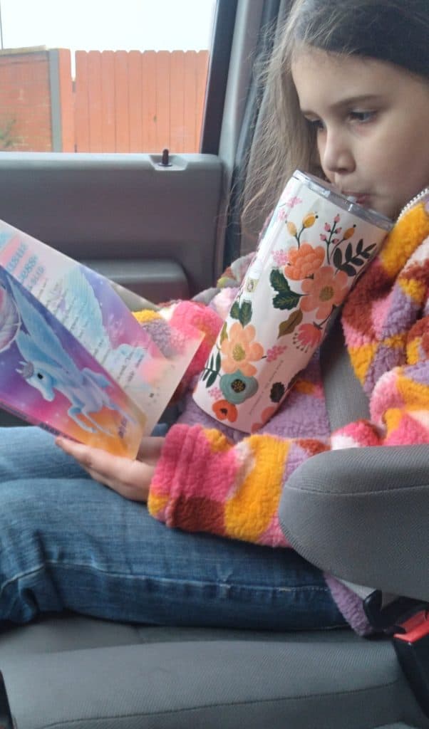 Young girl sitting in the car, drinking out of a floral Tumbler while reading a book.