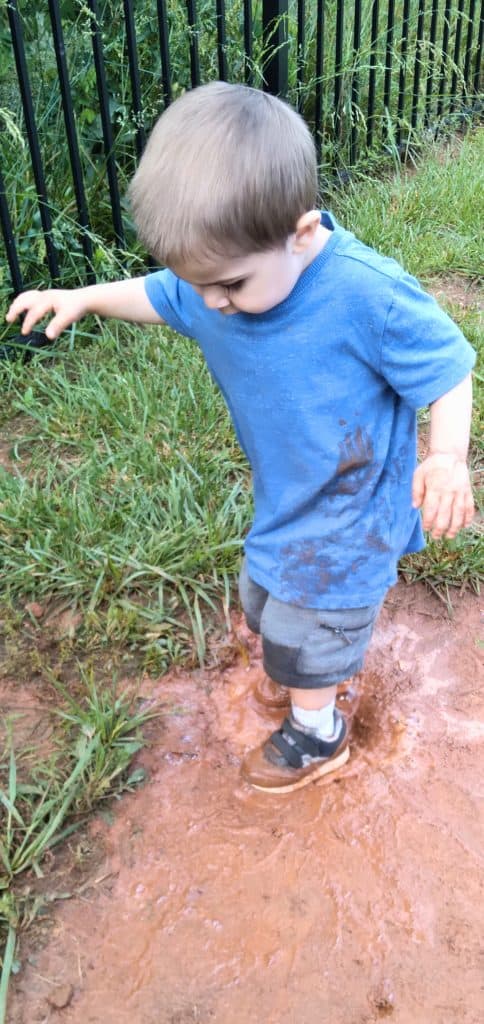 Little Boy wearing a blue shirt, grey shirt, and grey tennis shoes stomping in a mud puddle outside.