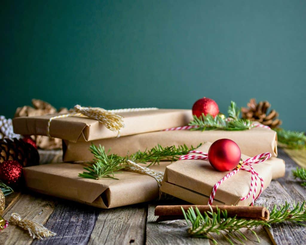 Four gifts wrapped in craft paper with pine needles, pines cones, and red ornament balls stacked around.