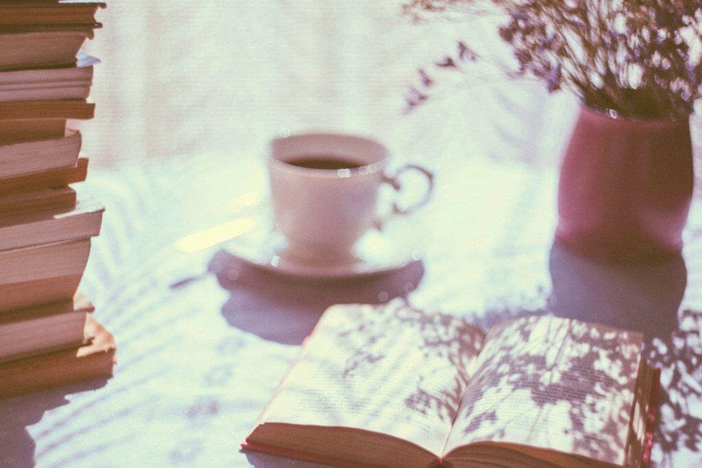White cup of tea beside a stack of books, vase of flowers, and open book on the table.