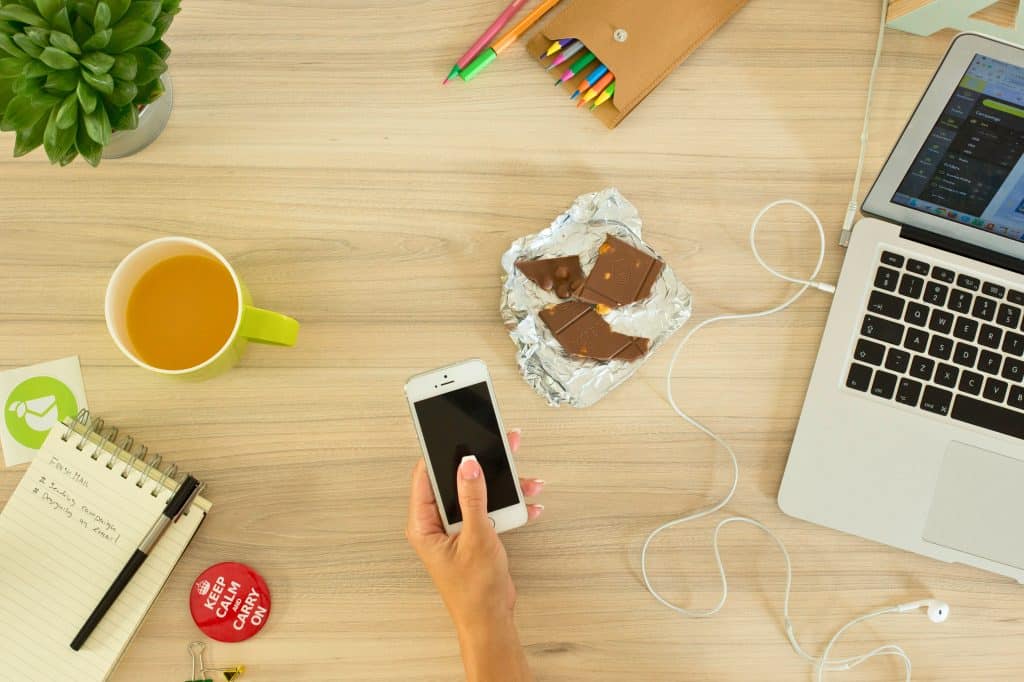 Cluttered table containing a laptop, colored pencils, broken up chocolate bar, cell phone, cup of tea, to do list, and a small plant.