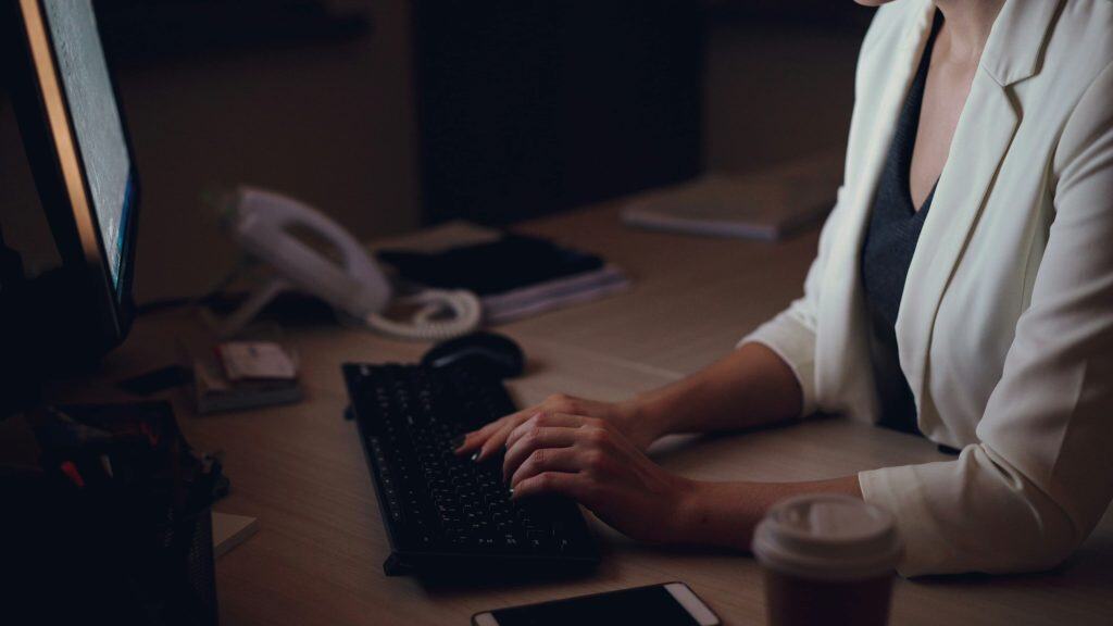 Woman wearing a black blouse and white blazer sitting down at her desk typing at her black keyboard.