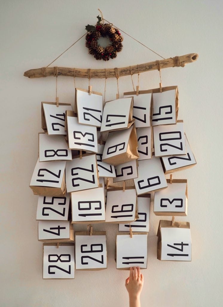 A small pinecone wreath hangs on a white wall above a homemade advent calendar made from a large stick and 25 brown paper bags with numbers on them hanging down from twine. A child's hand reaches for a bag.