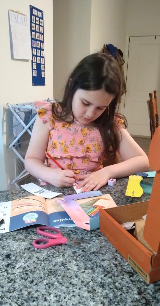 Young girl in a pink dress with yellow sunflowers sitting at the counter with a craft box open, scissors, paper, and colored pencils scattered around while she is coloring.