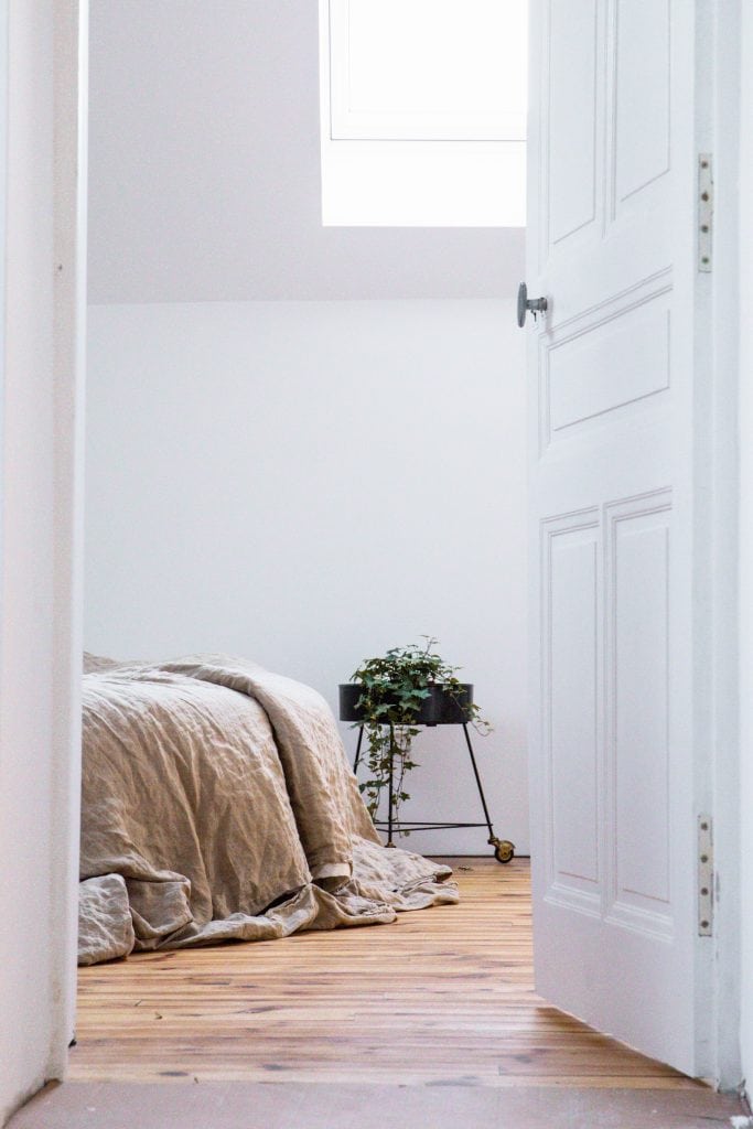 White bedroom door opened to a view of the foot of a bed covered in a tan linen comforter. A green potted plant sits on a black rolling cart beside the bed.