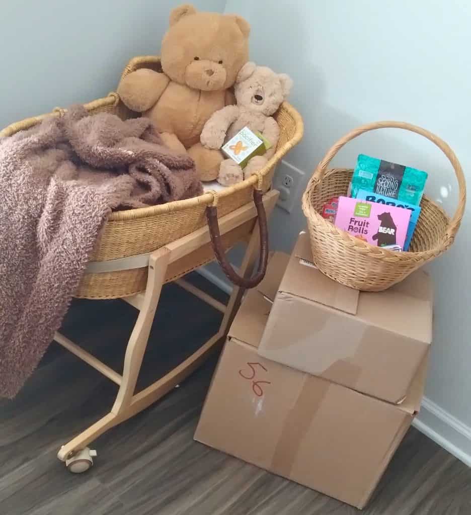 Wicker Bassinet in a wooden stand with various brown colored bears of various sizes sitting inside. A pecan brown blanket it tossed over the side. Beside that is a brown wicker basket filled with miscellaneous snacks stacked on top of a medium brown box and large brown box which contain various home birth kit supplies.