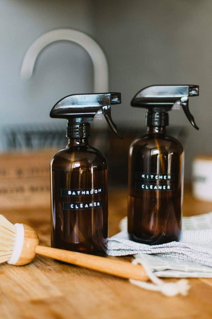 Two amber spray bottles with black labels that read Bathroom Cleaner and Kitchen Cleaner sitting beside a white and blue dishcloth and long handled wooden scrubber.
